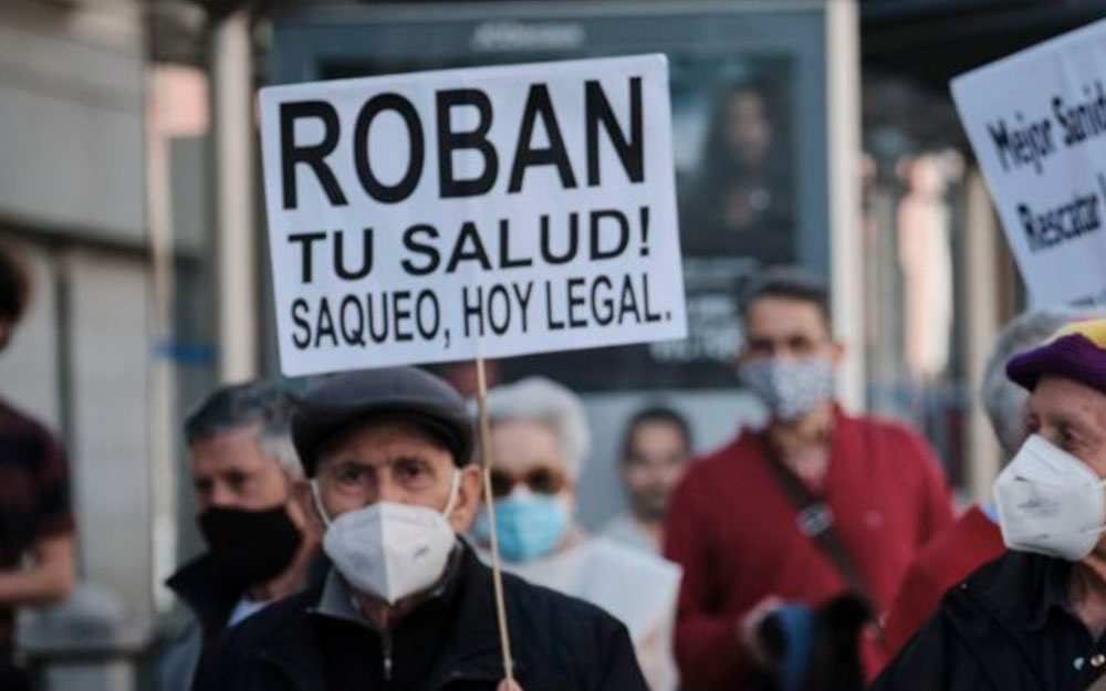Una persona mayor en una manifestación porta un cartel que dice "roban tu salud, saqueo hoy legal".
