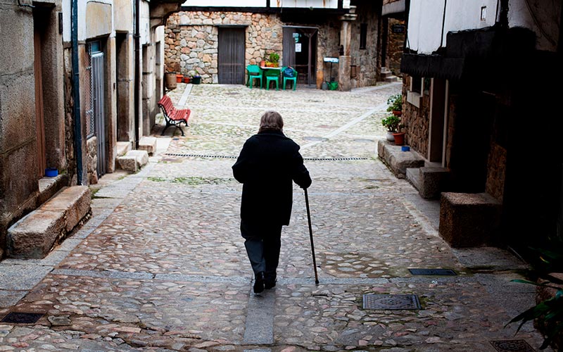 Una mujer mayor con un bastón camina sola por la calle de un pueblo 