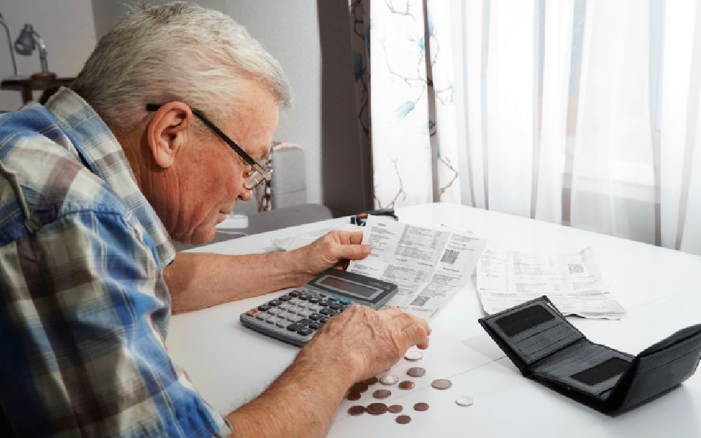 Un hombre mayor, con una calculadora, cuenta monedas sobre una mesa 