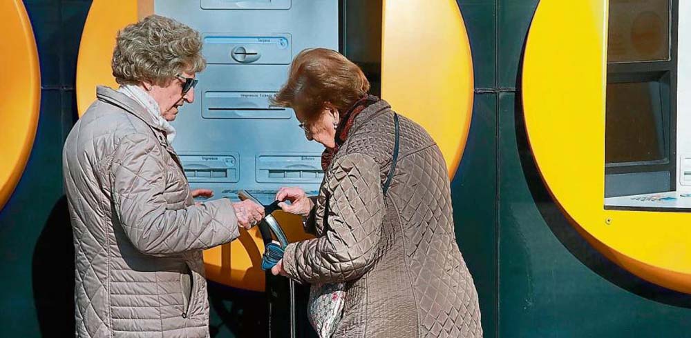 Una mujer mayor, ante un cajero, entrega a otra una libreta bancaria
