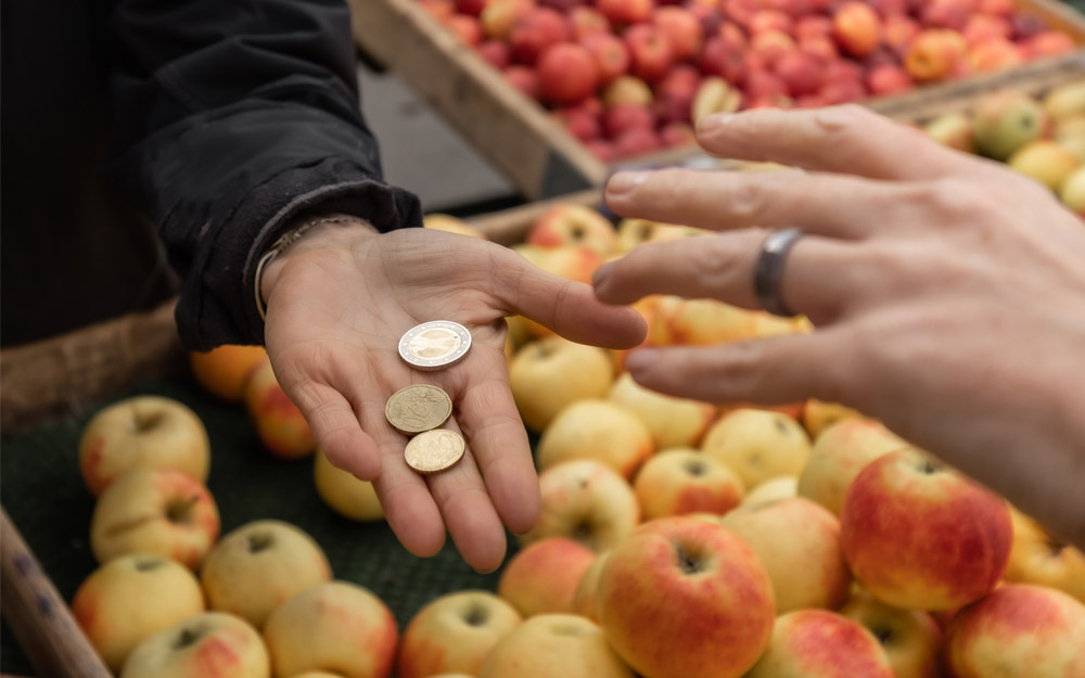 Una persona paga su compra en una frutería. En la imagen, se ven dos manos: una de ellas, con unas monedas; y la otra, a punto de cogerlas.