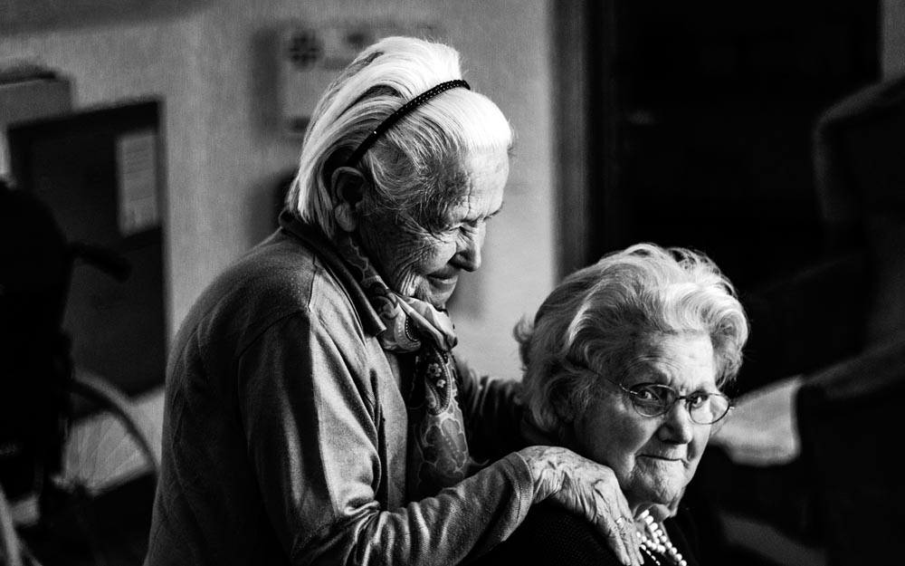 Las mujeres mayores sufren una doble discriminación: por edad y género. Dos mujeres mayores. Una de ellas aparece sentada y la otra, detrás, se apoya sobre sus hombros.