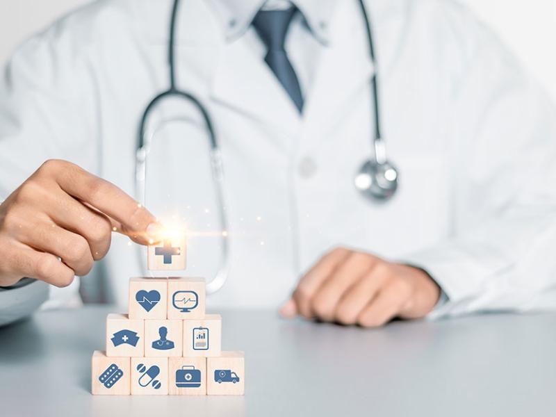 Una persona con bata de médico monta una pirámide de bloques de madera con iconos relacionados con la salud